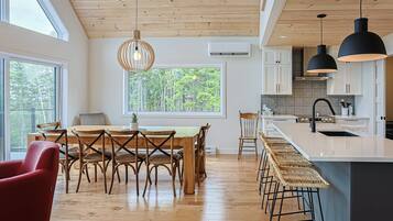 Una cocina moderna con pisos de madera, un comedor con una mesa y sillas de madera, y un amplio ventanal con vistas a la vegetación.