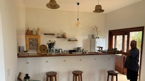 Eine Küche mit Holzstühlen, einem Esstisch mit grüner Tischdecke und einer Person am Arbeitsplatz.