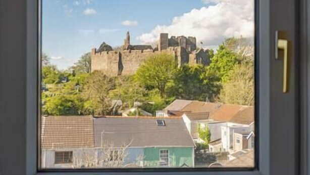 Visão de um castelo medieval numa colina, com vegetação e edifícios residenciais ao redor.