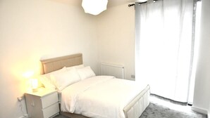 Ein Schlafzimmer mit Bett, Nachttisch, Lampe, Heizkörper und einem Fenster mit Vorhängen.