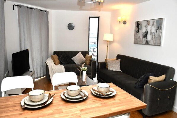 A modern living room with a black sofa, a wooden dining table with bowls and chopsticks, a TV, and a painting on the wall.