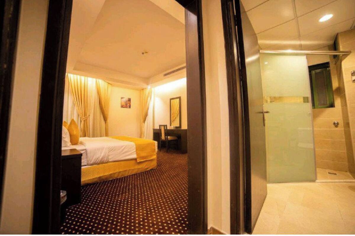 A hotel room with a bed, a desk, a chair, a bathroom with a glass door, and a patterned carpet.