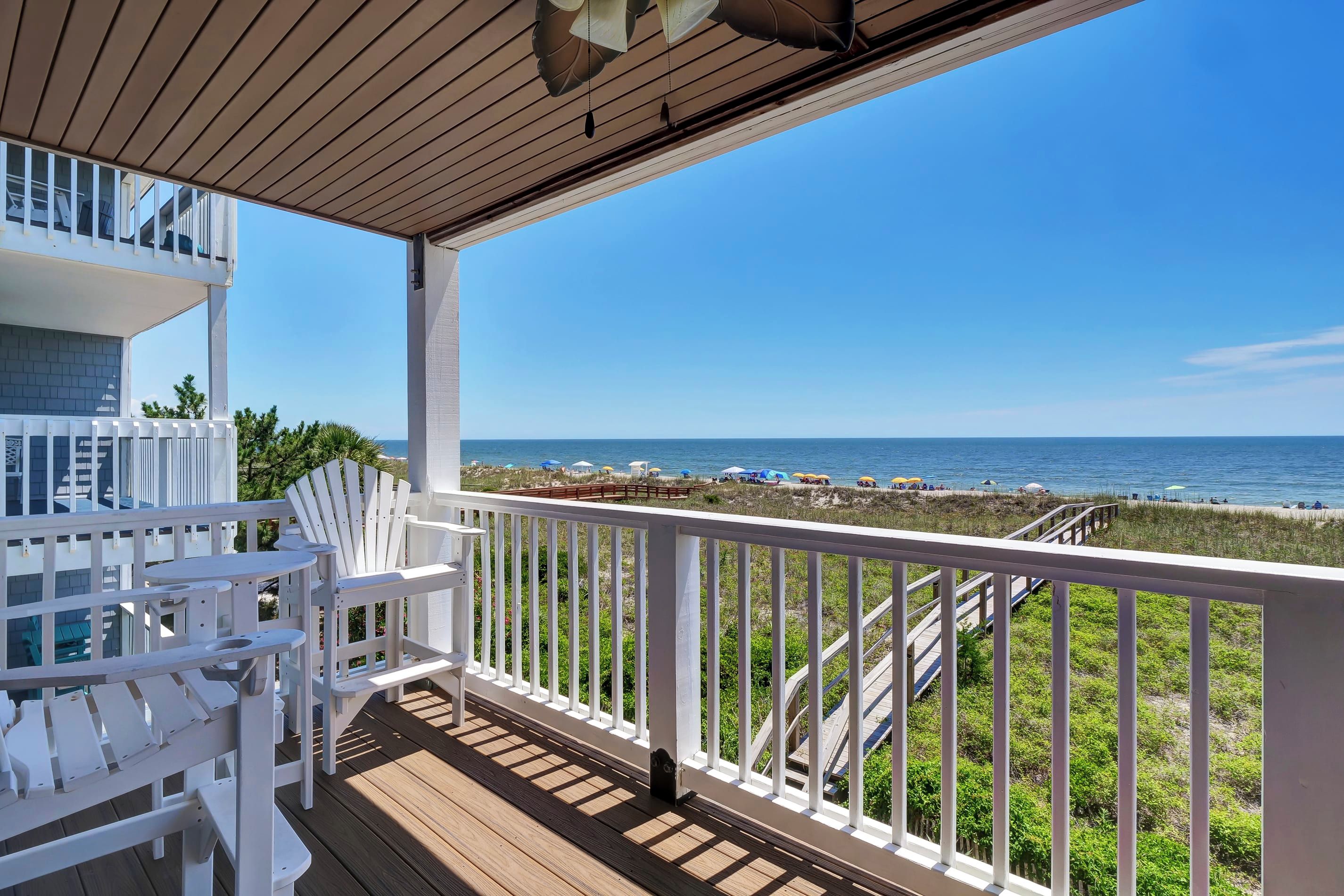 A wooden deck with white railings overlooking a beach with umbrellas and people.