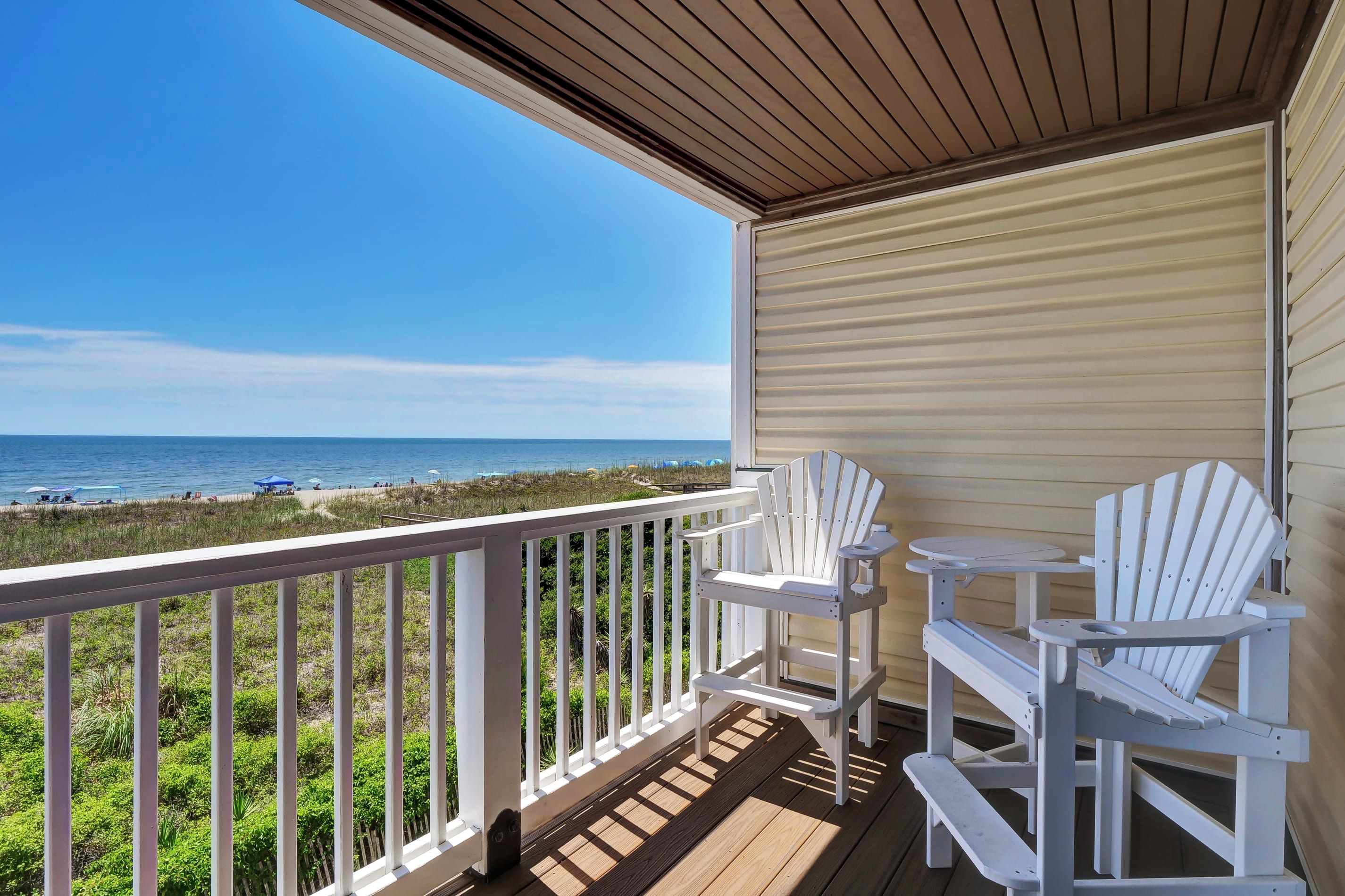 A covered deck with white Adirondack chairs and a small table, overlooking a beach with a clear view of the ocean.
