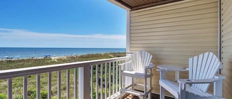 A covered deck with white Adirondack chairs and a small table, overlooking a beach with a clear view of the ocean.