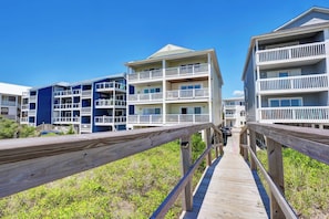 A wooden boardwalk leading to a beachfront building with multiple balconies and a clear blue sky.