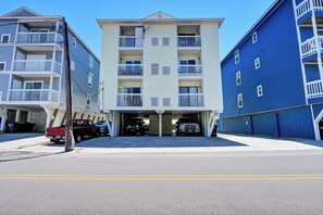 A multi-story building with a central entrance, balconies, and a parking area with vehicles.
