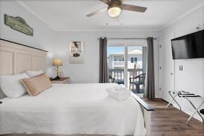 A bedroom with a bed, a ceiling fan, a TV, and a view of a house through the window.