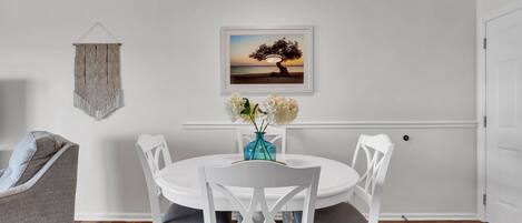 A dining area with a white table and chairs, a vase with flowers, and a framed picture on the wall.