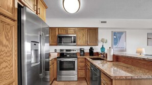 A modern kitchen with stainless steel appliances, wooden cabinets, a granite countertop, and a large island. - Infinity Summer, Oceanfront Retreat (Carolina Beach)