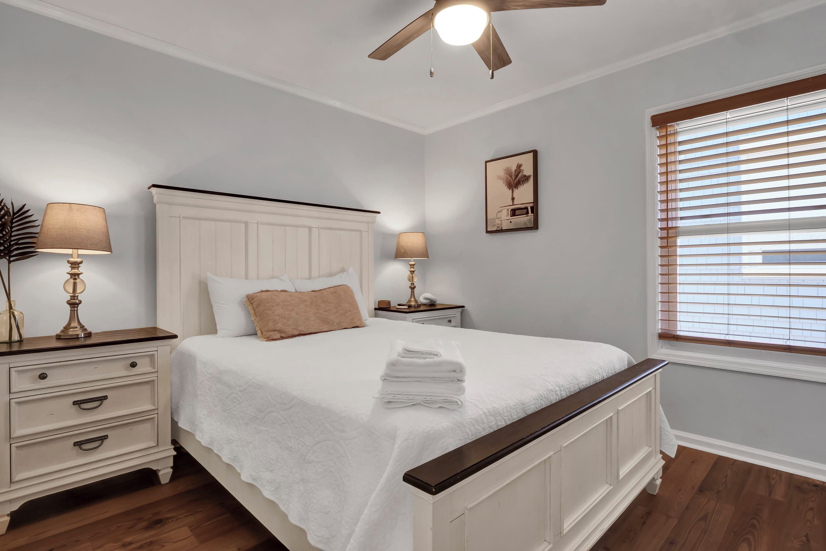 A bedroom with a bed, two bedside tables, a ceiling fan, a window with blinds, and a wall-mounted picture.