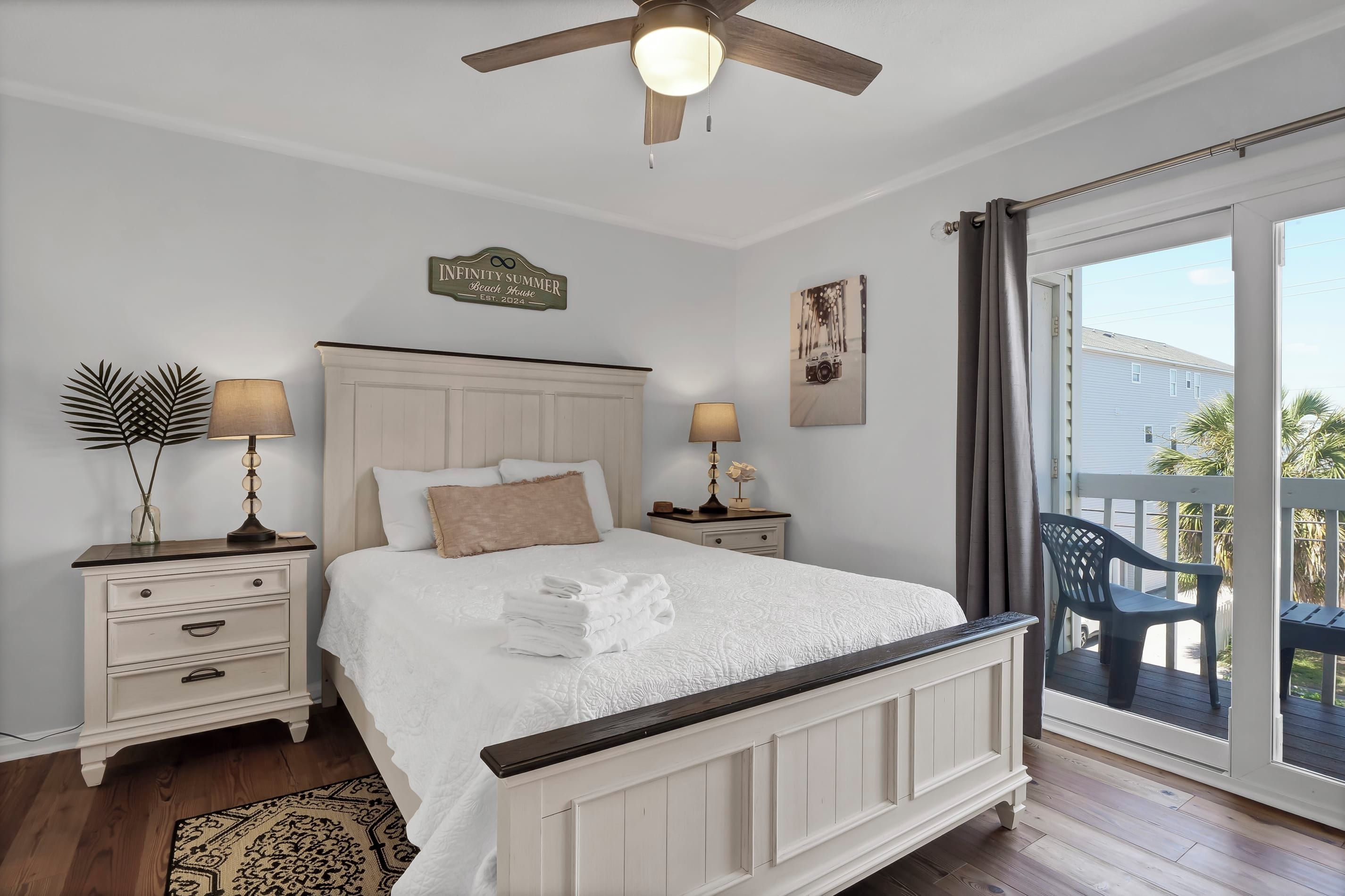 A bedroom with a bed, two bedside tables, a ceiling fan, a nightstand with a lamp, a wall clock, and a window with a view of the outside.