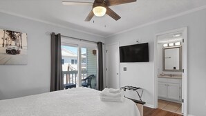 A bedroom with a bed, a ceiling fan, a TV, a bathroom with a mirror, and a balcony with a chair. - Infinity Summer, Oceanfront Retreat (Carolina Beach)