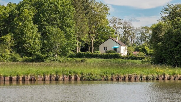 A house by a calm river with a grassy bank and dense trees in the background.