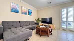 Un salon avec un canapé d’angle gris, un téléviseur à écran plat posé sur un meuble en bois, et trois tableaux représentant des paysages, encadrés et accrochés au mur.