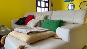 A living room with a beige sofa, multiple cushions, a small side table, and a window with blinds. - Apartamento Para 4 Personas en Playa Jardín Boiro, Puerto de Escarabote (Boiro)