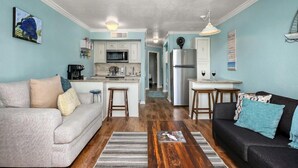 Una sala moderna con un sofá, una mesa de centro y un sofá negro. Se ve una cocina con armarios blancos y un refrigerador al fondo.