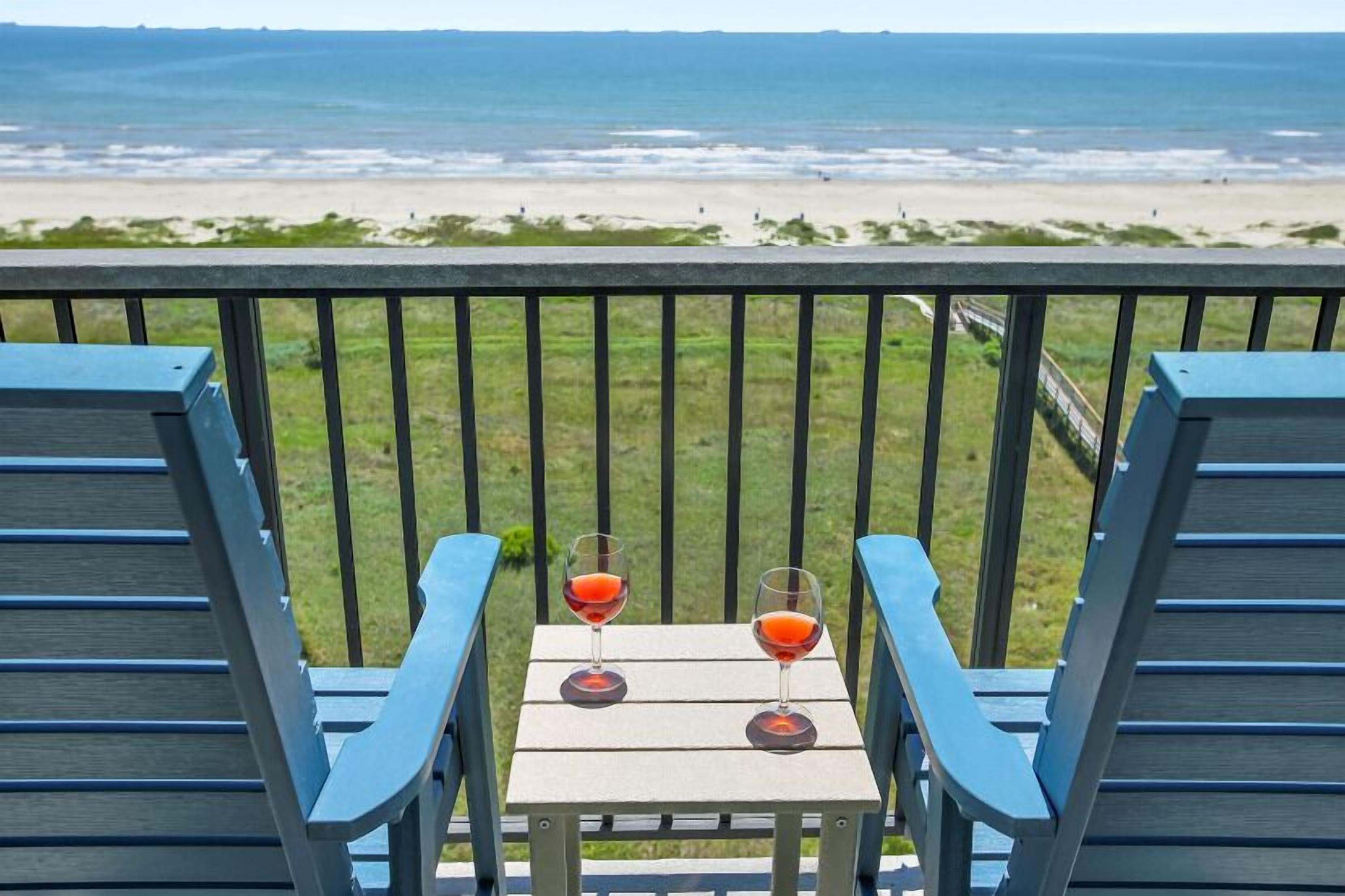 A balcony with two blue chairs and a small table, each with a glass of red drink. The view includes a beach and ocean.