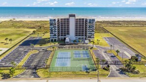 Een hotel met meerdere verdiepingen, een tennisbaan en een parkeerplaats, nabij een strand.