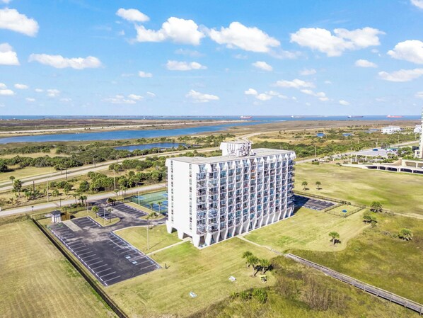 A high-rise hotel with multiple balconies, a parking lot, and a view of a body of water and greenery.