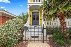 A yellow house with a grey door, surrounded by a black metal fence, and a palm tree to the right.