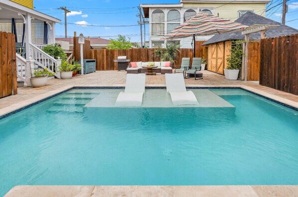 A backyard with a swimming pool, a wooden deck, a covered seating area, and a fenced yard.