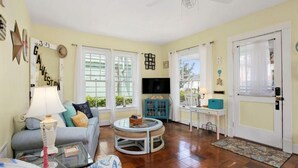 A living room with a sofa, coffee table, and TV. There are windows, a ceiling fan, and a door with a curtain. - Welcome to "The Sunrise Inn!" (Galveston)