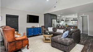 Ein modernes Wohnzimmer mit Ledersessel, grauem Sofa mit Kissen und einem blauen Schrank, im Hintergrund eine Küche.
