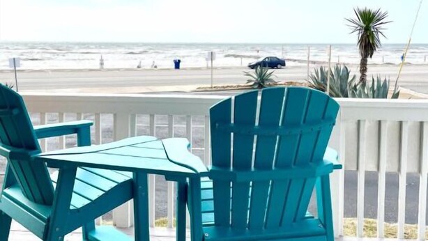 A turquoise deck with two chairs and a table, overlooking a beach with a fence and palm trees.