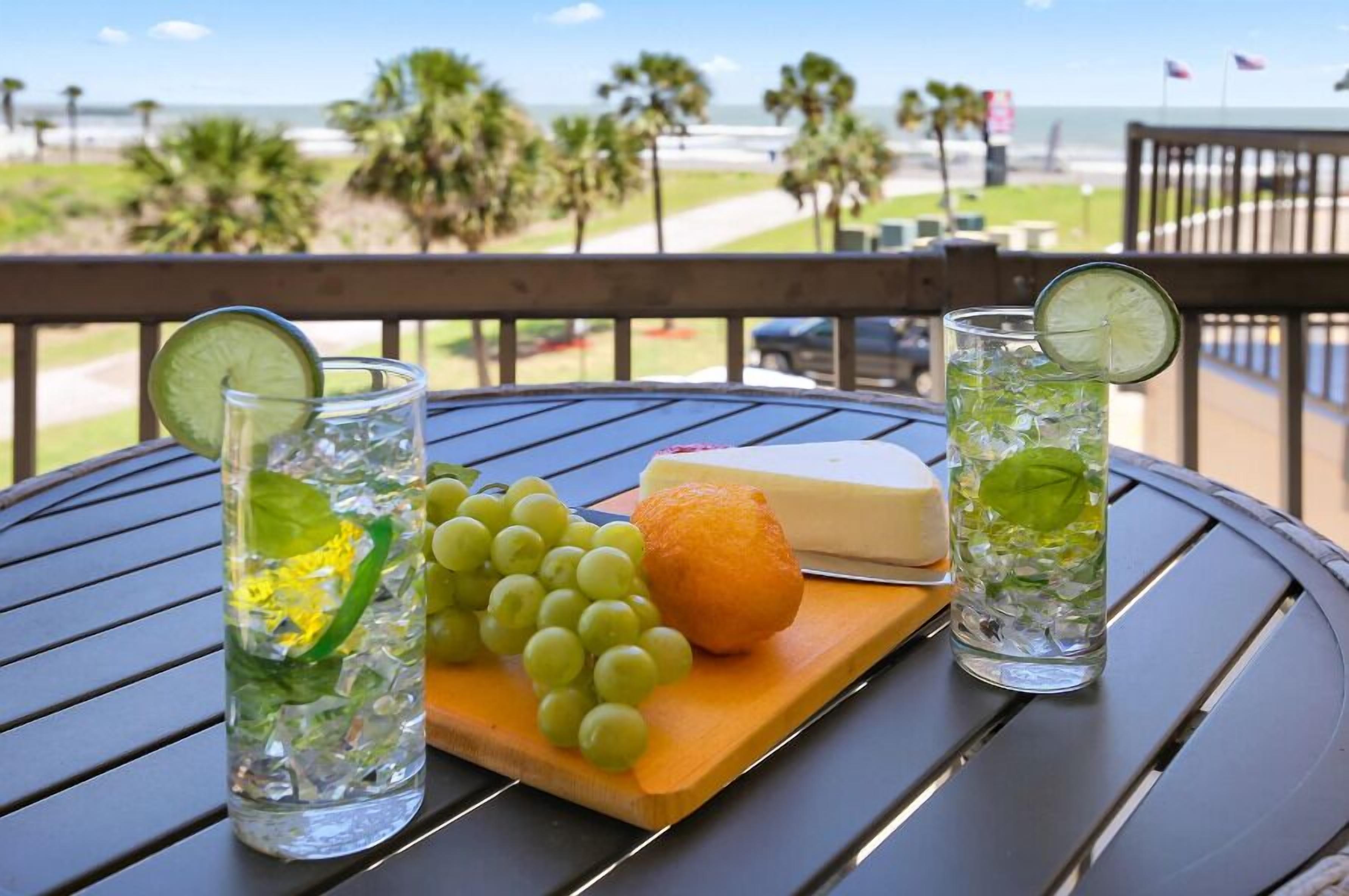 Un tavolo con due bicchieri di bevande fredde, formaggio, acini di uva e una fetta di limone su un tagliere, con vista sulla spiaggia e palme sullo sfondo.