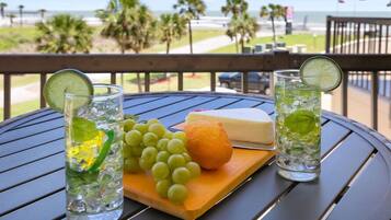 Una mesa con dos vasos de bebidas heladas, queso, uvas y una rodaja de limón sobre una tabla de madera, con vista a la playa y palmeras al fondo.