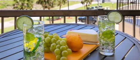 Une table avec deux verres de boissons glacées, un morceau de fromage, des raisins et une tranche de citron sur une planche en bois, avec en arrière-plan une vue sur la plage et des palmiers.