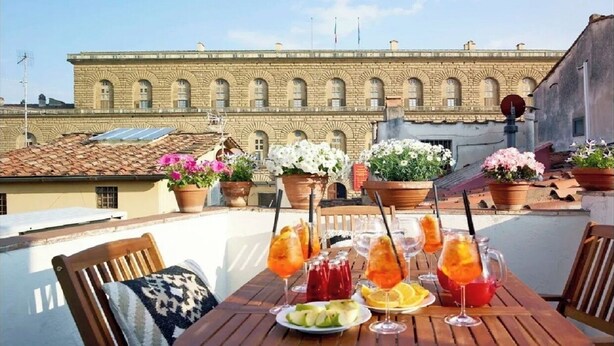 Una terraza en la azotea con una mesa lista para comer, plantas en macetas y un edificio con ventanas de arco en el fondo.