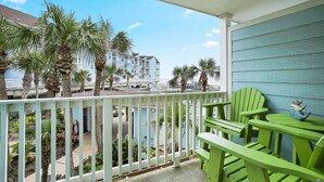 A balcony with green Adirondack chairs, a small table, and a view of a pool and buildings in the background.