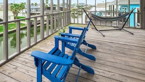 A covered deck with blue Adirondack chairs and a hammock, overlooking a waterway with buildings and palm trees. - Sea Isle Sailfish! Canal front in popular Sea Isle! Pet Friendly! (Galveston)