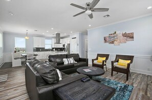 Una sala de estar moderna con un sofá, una mesa de centro y dos sillones. Se ve una cocina al fondo.