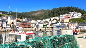 A coastal town with buildings along the waterfront, a fishing boat docked, and a hill in the background. - The Cliffs - ViveMare (Viveiro)