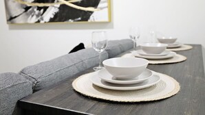A dining table set with white bowls and glasses on woven placemats, against a grey sofa and a black and white abstract painting. - Elegant Escape w/King Bed|Long Stays|WEM|Disney+ (Edmonton)