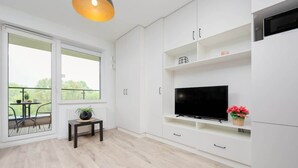 A modern living room with a TV on a white cabinet, a small table with a plant, and a balcony with a table and chairs. - Comfortable Studio with Balcony | Close to Metro and Park (Warszawa)