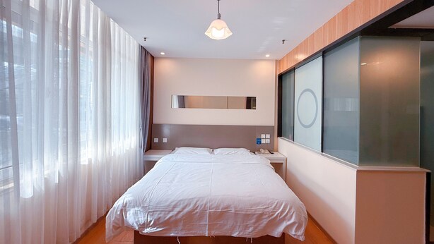 A modern bedroom with a large bed, a bedside table, a mirror, and a window with curtains. - Morning Wind Urban Hotel （May Fourth Square Branch) (Qingdao)