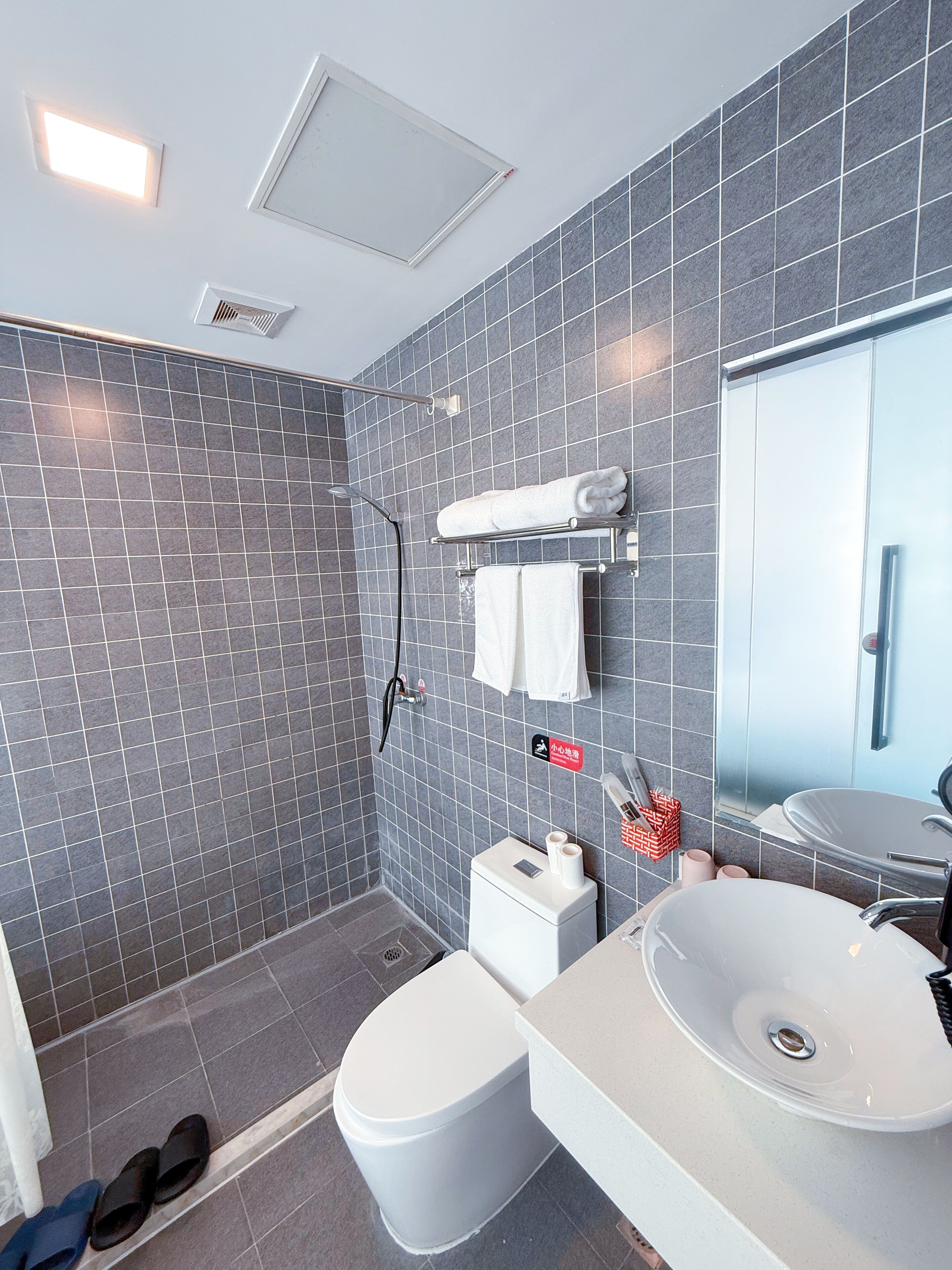 Un baño moderno con paredes de azulejos grises, un inodoro blanco, una ducha y un lavabo con espejo.