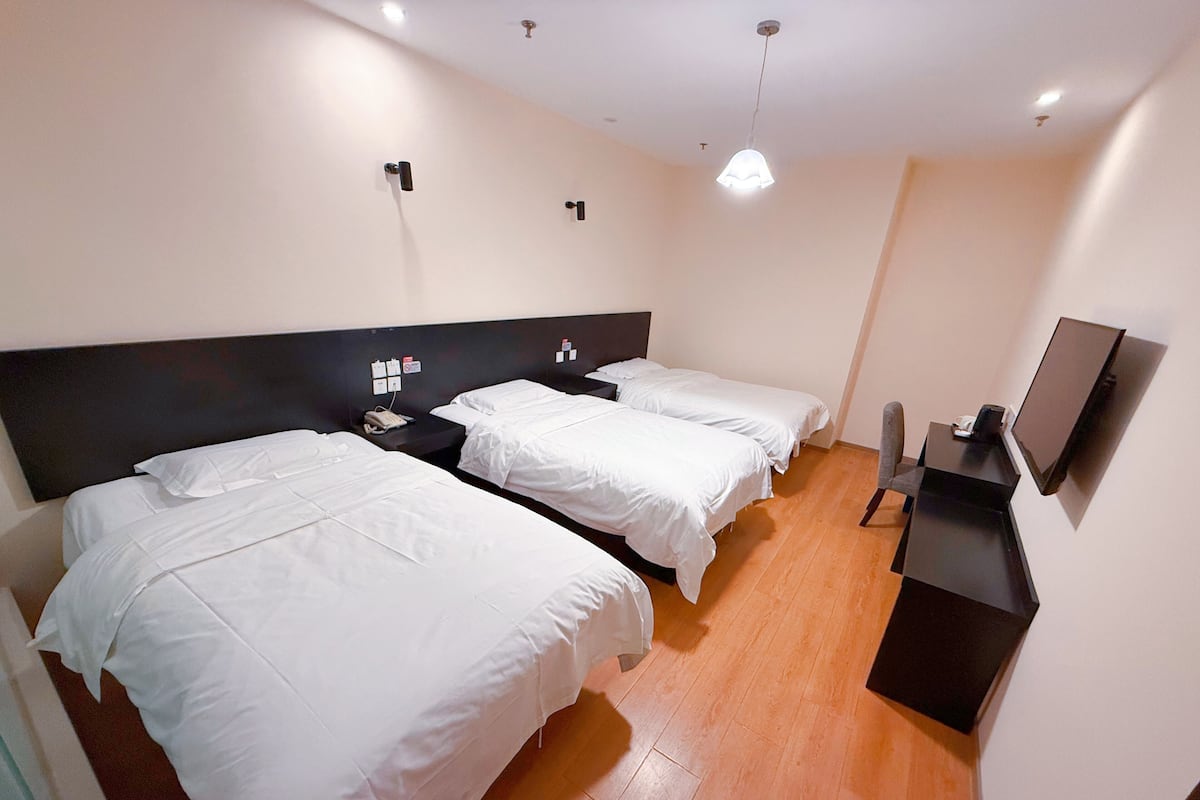 Ein Hotelzimmer mit drei Einzelbetten, einem Schreibtisch, einem Stuhl, einem Fernseher und einer Deckenleuchte.