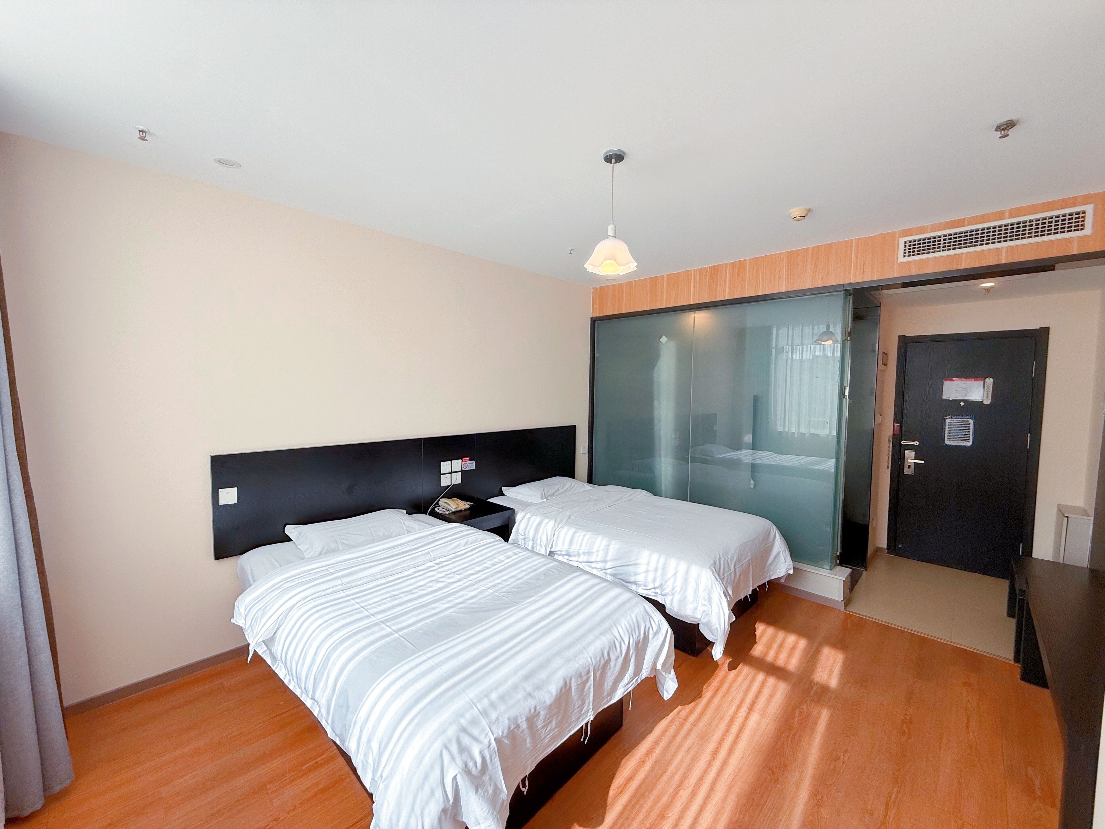 A modern hotel room with two beds, a wooden floor, a black headboard, a ceiling light, and a sliding glass door.