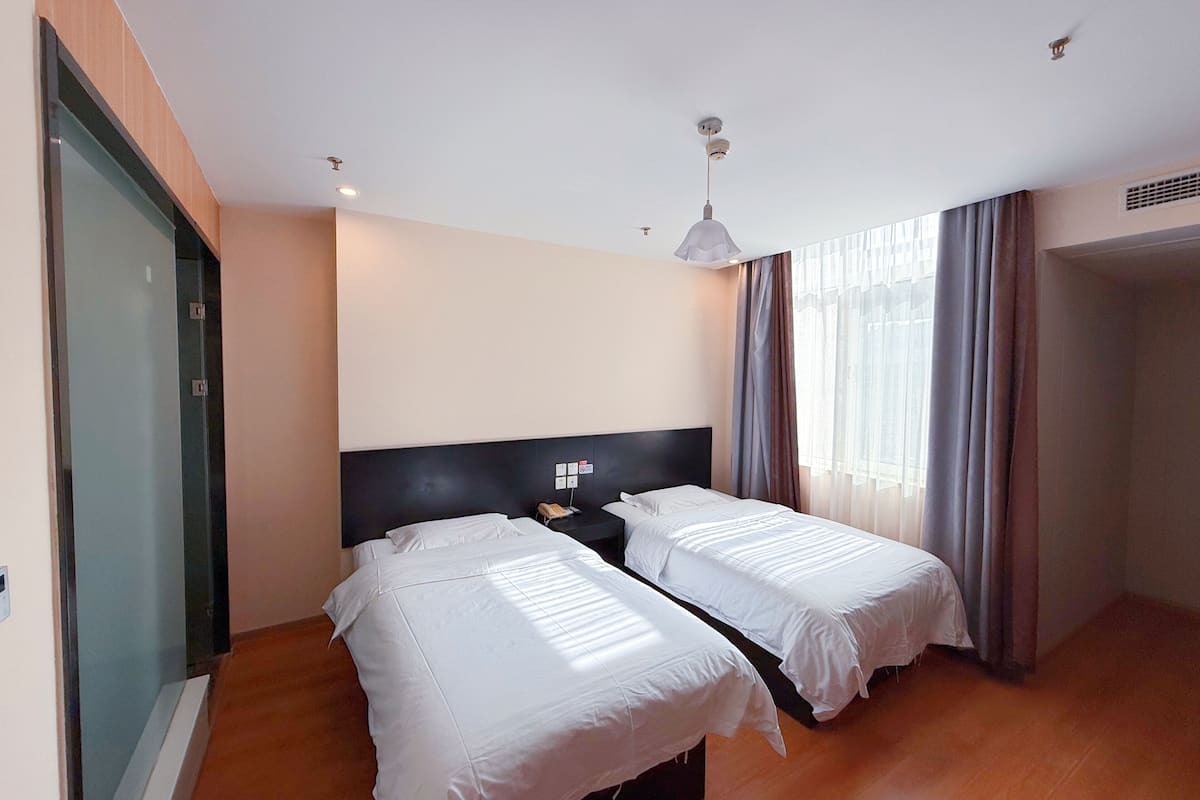 Ein Hotelzimmer mit zwei Betten, einem dunklen Kopfteil, einem Nachttisch, einer Pendelleuchte und einem großen Fenster mit Vorhängen.