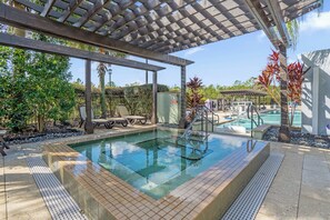 Ein Schwimmbecken mit einer hölzernen Pergola darüber, umgeben von tropischen Pflanzen und blauem Himmel.