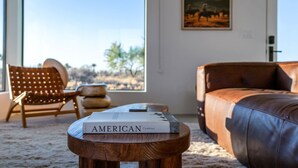 Una habitación con un sofá de cuero, una mesa de centro de madera con un libro, una silla de mimbre y un ventanal con vistas al desierto.