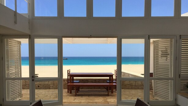 A room with a view of a beach through large windows. There is a wooden bench and a table with chairs. - 4 Bedroom Private vacation home in Vila do Maio (Vila do Maio)