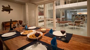 A dining area with a wooden table set for a meal, a view of the kitchen, and a large window with blinds. - 4 Bedroom Private vacation home in Vila do Maio (Vila do Maio)