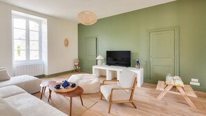 Ein Wohnzimmer mit grüner Wand, einem weißen Sofa, einem Holztisch, einem weißen Sessel, einem Fernseher und einem kleinen Holztisch mit Gegenständen darauf.