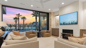 A modern living room with a large window overlooking a pool and palm trees, a fireplace, and a mounted TV.